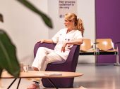 Blonde vrouw in witte verpleegkundige kleding zit op een paarse stoel. ze kijkt naar links waar ze in gesprek is met iemand die wij niet zien.
