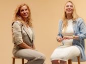Twee lachende vrouwen zitten naast elkaar op krukken voor een beige achtergrond.