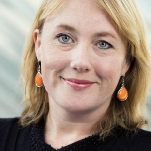 Vrouw met blond haar en oranje oorbellen kijkt glimlachend in de camera.