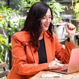 Vrouw in oranje jasje zit achter laptop met groene planten achtergrond