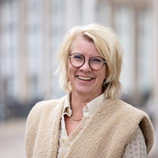 Blonde vrouw in beige kleding met bril lacht naar camera