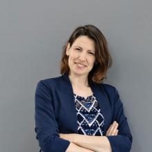 vrouw met halflang bruin haar staat met haar armen over elkaar heen voor een grijze achtergrond