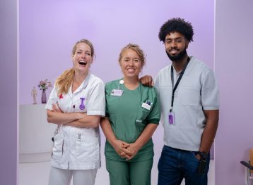 2 blonde vrouwen en een man met zwart haar en baard. Ze staan voor een paarse achtergrond en lachen. De eerste vrouw lacht voluit.
