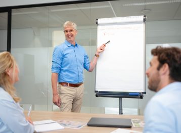 Man met blauw overhemd staat voor flipover en wijst er met een viltstift naar. een man en vrouw kijken naar hem.