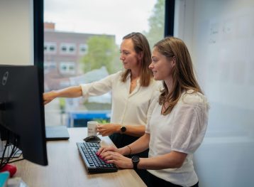 Twee vrouwen met donker haar en witte shirts kijken gezamelijk naar een computerscherm. we zien ze van de linkerkant, op de achtergrond een raam.