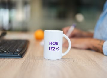 Op een bureau staat een koffiebeker met de tekst 'Hoe IZZ’t', met een sinaasappel en een toetsenbord op de achtergrond.