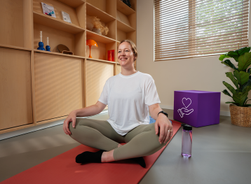 Blonde vrouw, zittend in kleermakerszit op yoga mat met paars blok met illustratie met hand en hartje