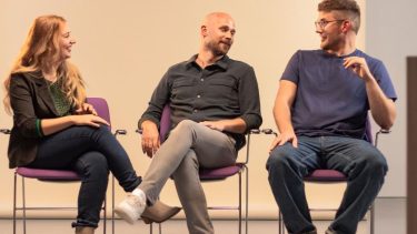 Vrouw en 2 mannen zitten naast elkaar op paarse stoelen en praten met elkaar