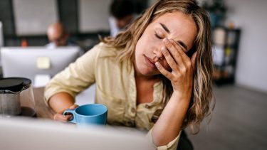 Een vrouw met blond haar zit achter haar computer en houdt vermoeid haar hand voor haar ogen.