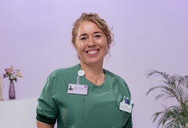 lachende blonde vrouw in groene operatiekleding staat voor een paarse achtergrond. Ze heeft 1 hand in de zak van haar shirt