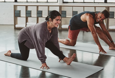 Focus ligt op lachende vrouw met donkerhaar in grijze yogakleren op yogamat. Ze steunt op haar handen en steekt 1 been naar voren. Op de achtergrond een man in dezelfde pose.