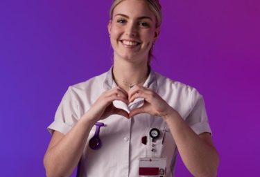 Lachende zorgmedewerker in uniform maakt hartgebaar met handen voor paarse achtergrond.