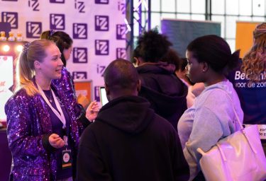 Meerdere mensen zijn met elkaar in gesprek bij een stand op een beursvloer van IZZ.
