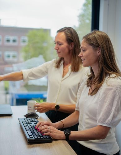 Twee vrouwen met donker haar en witte shirts kijken gezamelijk naar een computerscherm. we zien ze van de linkerkant, op de achtergrond een raam.