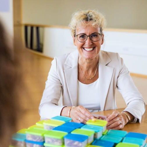 Vrouw met bril en krullend haar lacht terwijl ze aan tafel zit met stapels gekleurde doosjes.