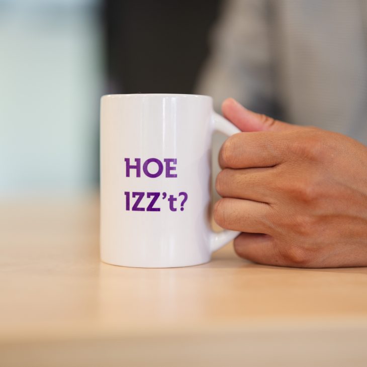 Hand houdt witte mok vast met paarse tekst: Hoe IZZ't?