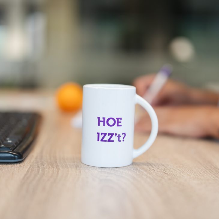 Op een bureau staat een koffiebeker met de tekst 'Hoe IZZ’t', met een sinaasappel en een toetsenbord op de achtergrond.