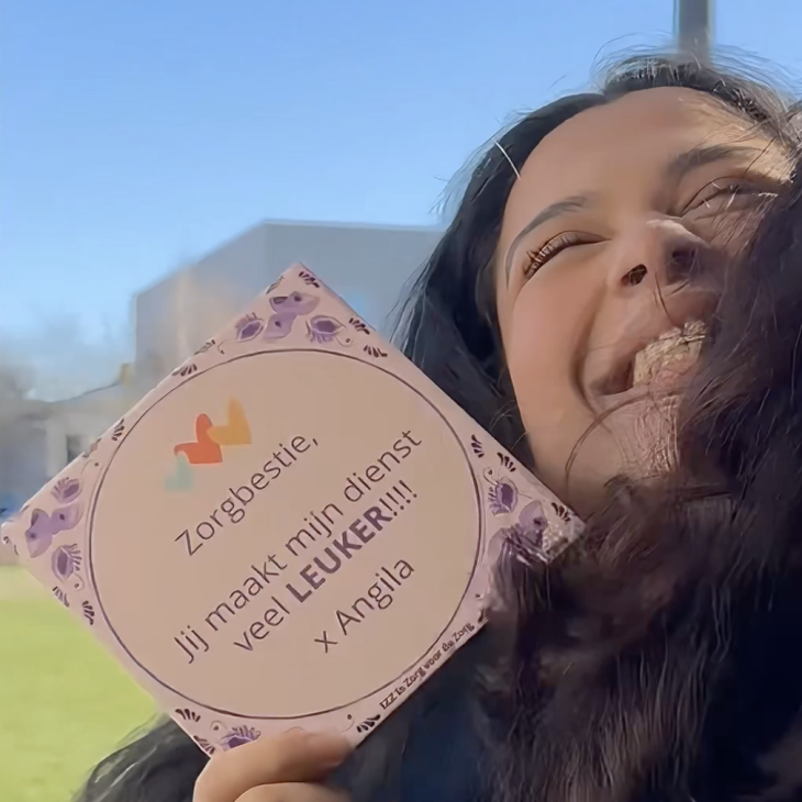 Een vrouw geeft een knuffel aan een ander en houdt in haar handen een paars Delfs blauw tegeltje waarop staat haar zorgbestie haar dienst veel leuker maakt.