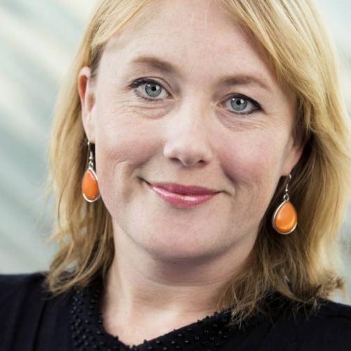 Vrouw met blond haar en oranje oorbellen kijkt glimlachend in de camera.