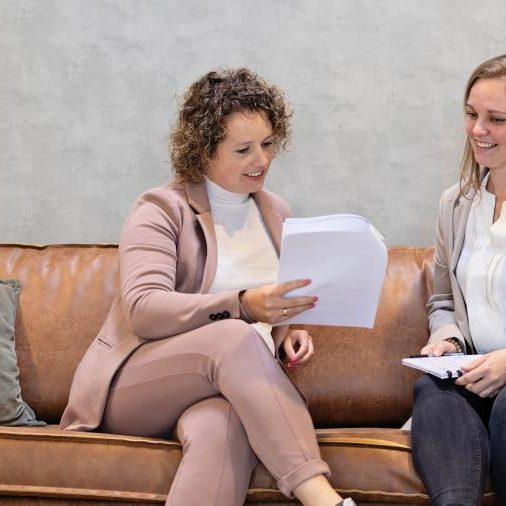 Twee vrouwen kijken naar papieren zittend op een bruine bank