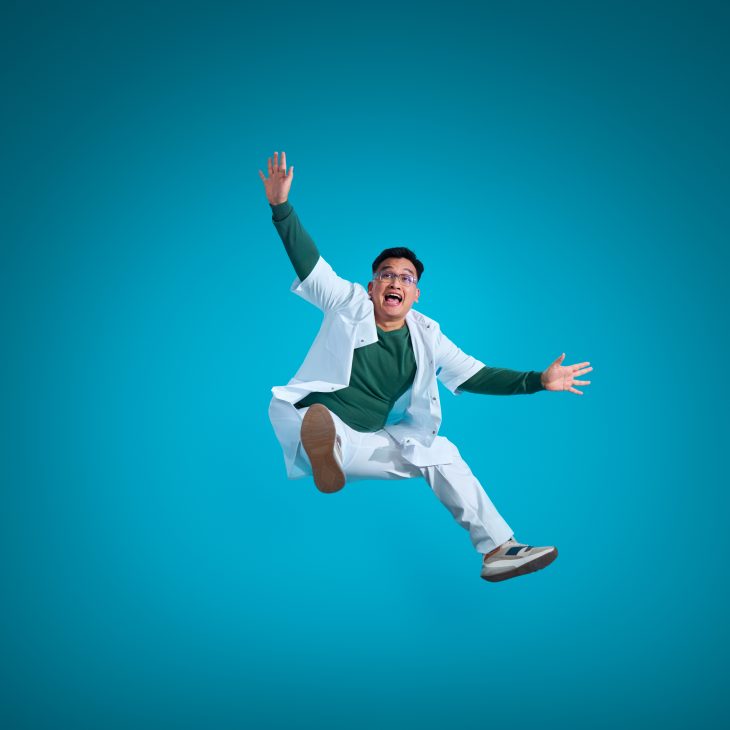 Springende man met zwart haar in een wit verplegingspak met een groen shirt eronder op een blauwe achtergrond