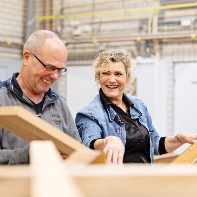 Blonde vrouw in spijkerjas lacht naar man in werkplaats