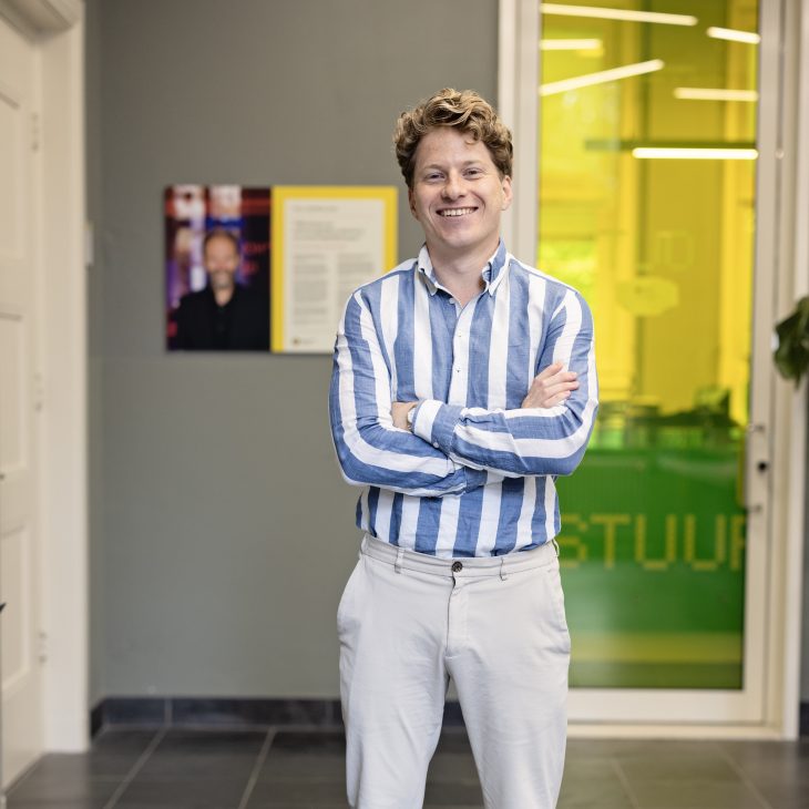 Staande man met een blauw wit gestreept overhemd voor een gele deur