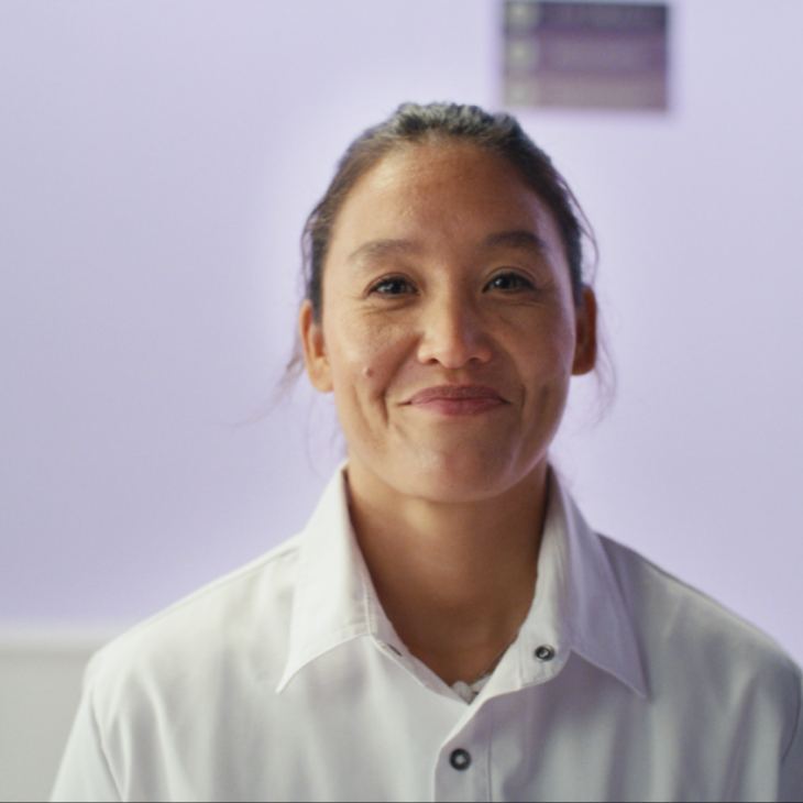Vrouw met donker haar in een staart kijkt naar voren. ze draagt een witte verpleegkundige jas.