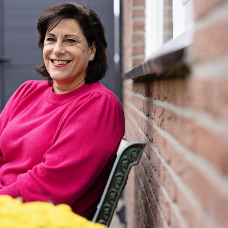 Een lachende vrouw in een roze trui zit op een ijzeren bankje tegen een bakstenen muur.