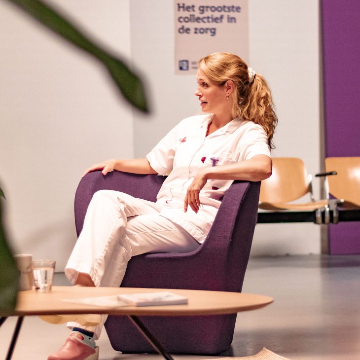 Blonde vrouw in witte verpleegkundige kleding zit op een paarse stoel. ze kijkt naar links waar ze in gesprek is met iemand die wij niet zien.