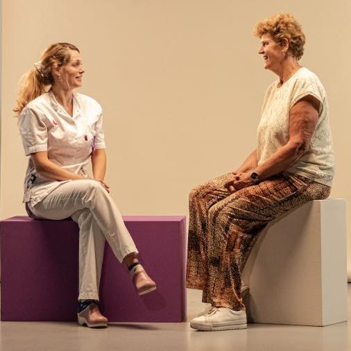 Zorgmedewerker en vrouw zitten tegenover elkaar op blokken en praten met elkaar in een studio.