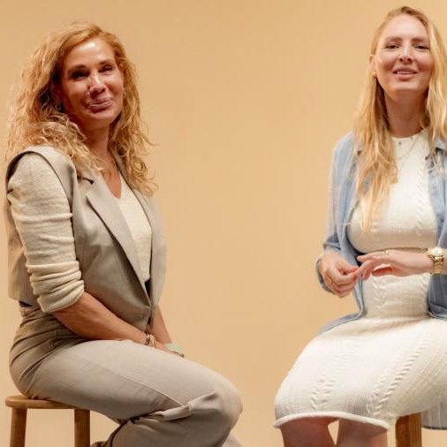 Twee lachende vrouwen zitten naast elkaar op krukken voor een beige achtergrond.