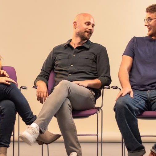 Vrouw en 2 mannen zitten naast elkaar op paarse stoelen en praten met elkaar
