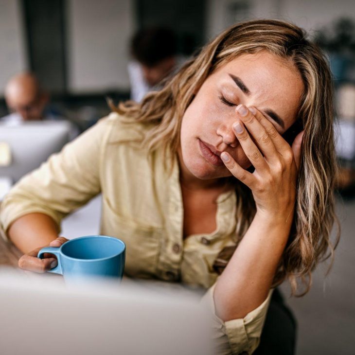 Een vrouw met blond haar zit achter haar computer en houdt vermoeid haar hand voor haar ogen.